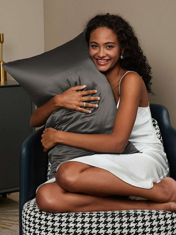 Smiling woman in white silk slip dress hugging a charcoal mulberry silk pillowcase.