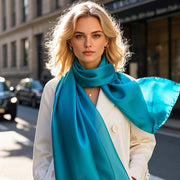 Blonde woman wearing a vibrant teal mulberry silk scarf/shawl over a white coat on a city street.