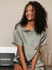 Smiling woman in a sage green mulberry silk short sleeve round neck pajama top with a chest pocket.