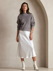Woman in a gray ribbed sweater and a white mulberry silk midi skirt with white boots.