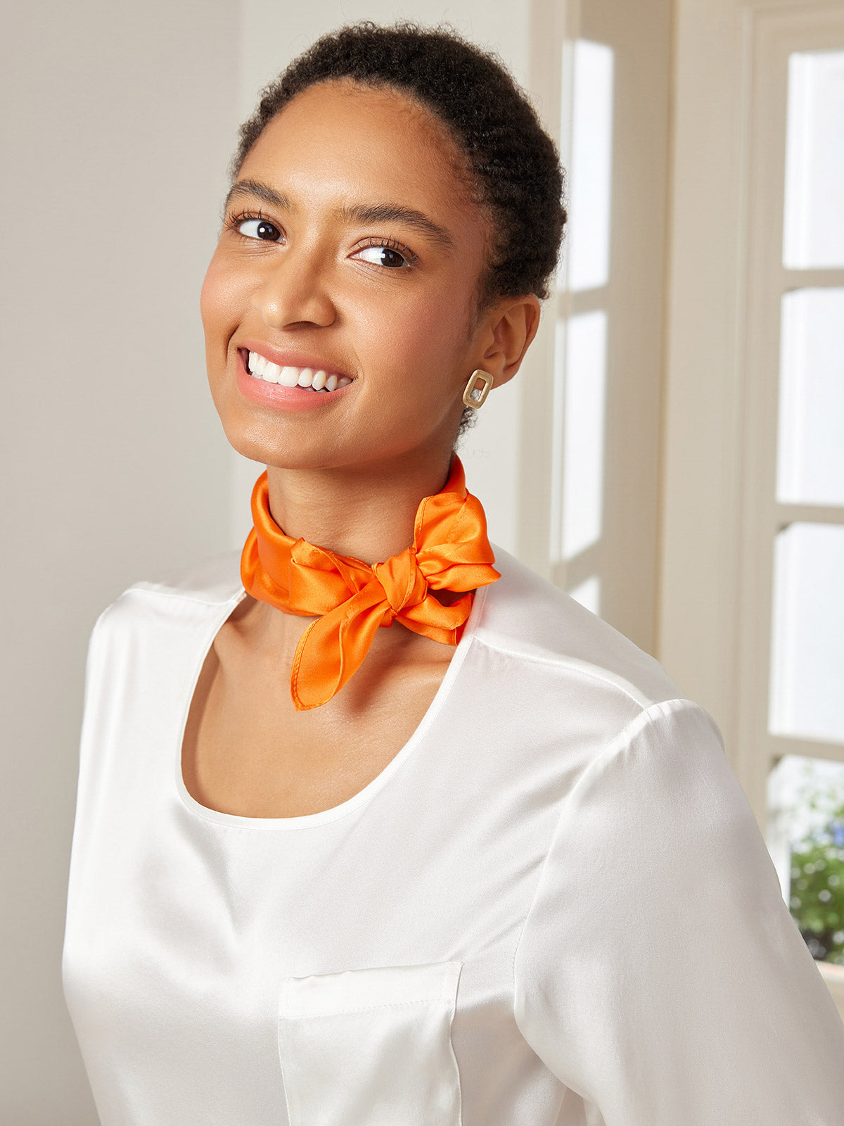 Woman wearing a vibrant orange mulberry silk square scarf tied around her neck, paired with a white silk top.