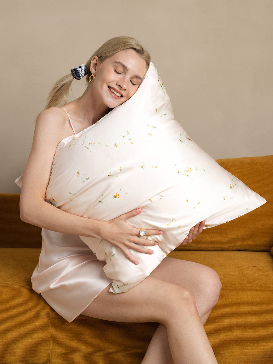 Blonde woman in silk slip hugging a Snowbud Lily 19Momme mulberry silk pillowcase with delicate floral pattern.