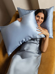 Smiling woman on a mustard sofa with two-tone light blue 19 Momme mulberry silk pillowcases.