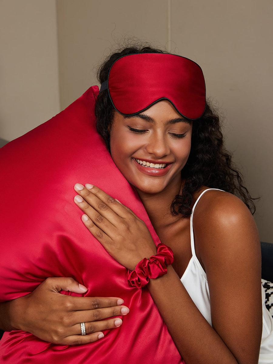 Smiling woman in white camisole hugging a wine red mulberry silk pillow with matching eye mask and scrunchie.