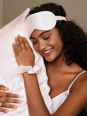 Smiling woman wearing a pink mulberry silk sleep mask and scrunchie, hugging a matching silk pillowcase.