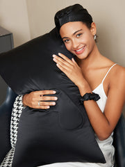 Smiling woman holding a black 19Momme mulberry silk pillowcase, wearing a matching silk bonnet, eye mask, and scrunchie.