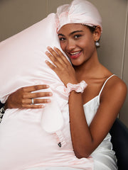 Smiling woman in a white silk camisole, hugging a pink mulberry silk pillow, wearing a matching silk bonnet, eye mask, and scrunchie.