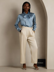 Women's 19Momme Mulberry silk long sleeve tie neck blouse in blue, paired with cream trousers.